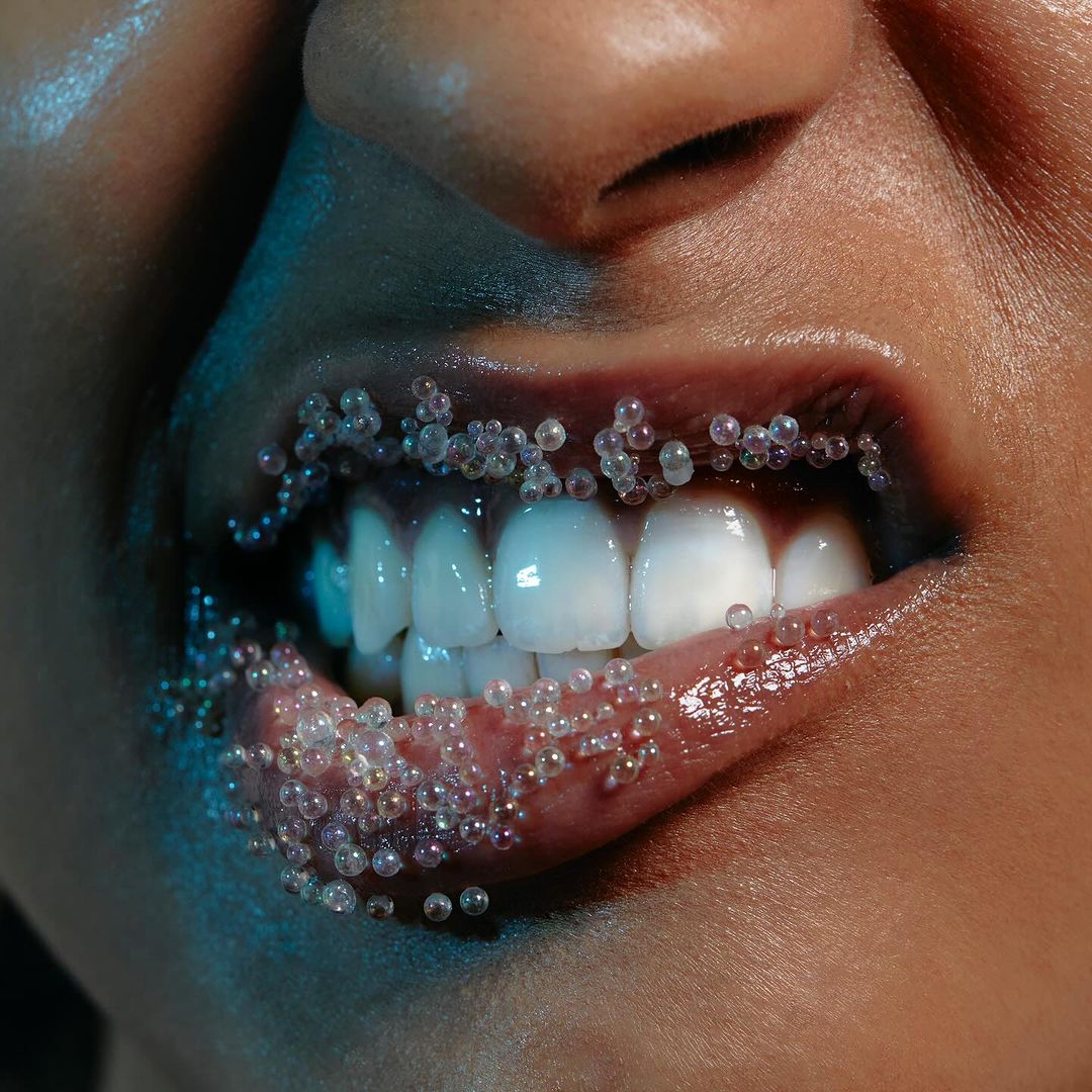 Close-up of a woman's lips adorned with translucent beads. The focus is on the teeth, lips, and beads under soft, bluish lighting.