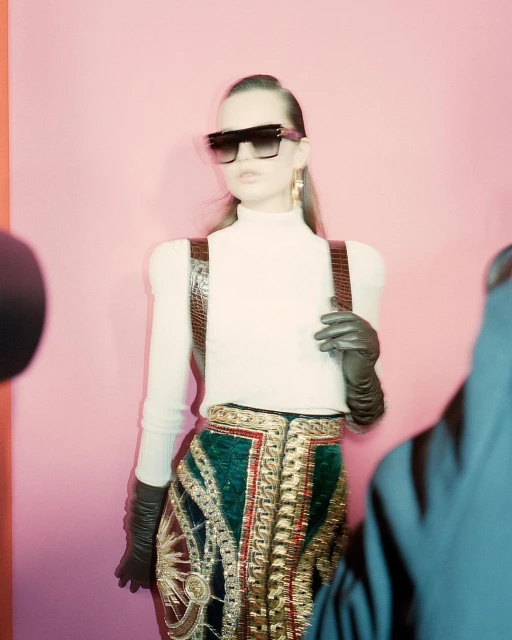 Fashionable woman in sunglasses and embellished skirt poses in a pink studio. Leather suspenders and gloves accessorize the outfit.