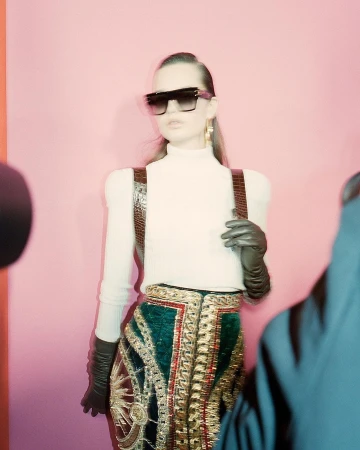 Fashionable woman in sunglasses and embellished skirt poses in a pink studio. Leather suspenders and gloves accessorize the outfit.