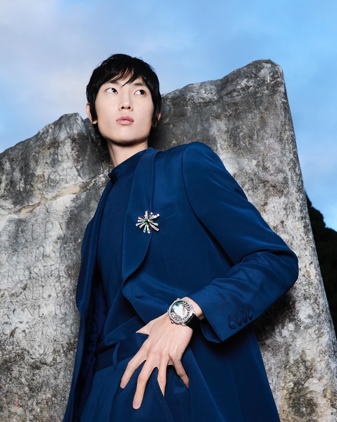 Adult male model in a blue suit poses near a weathered stone structure under a blue sky in an outdoor fashion shot.