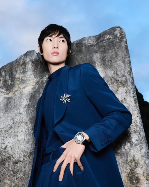 Adult male model in a blue suit poses near a weathered stone structure under a blue sky in an outdoor fashion shot.