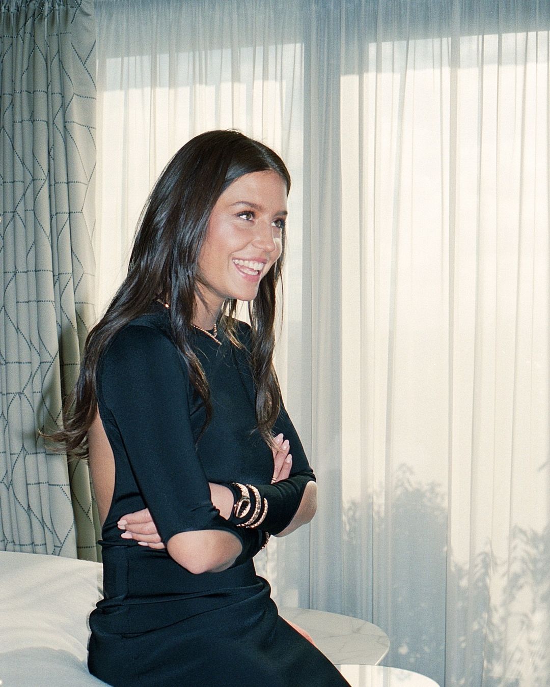 Brunette woman with a joyful expression poses indoors wearing a black dress and bracelets, with a window and patterned curtains in the background.