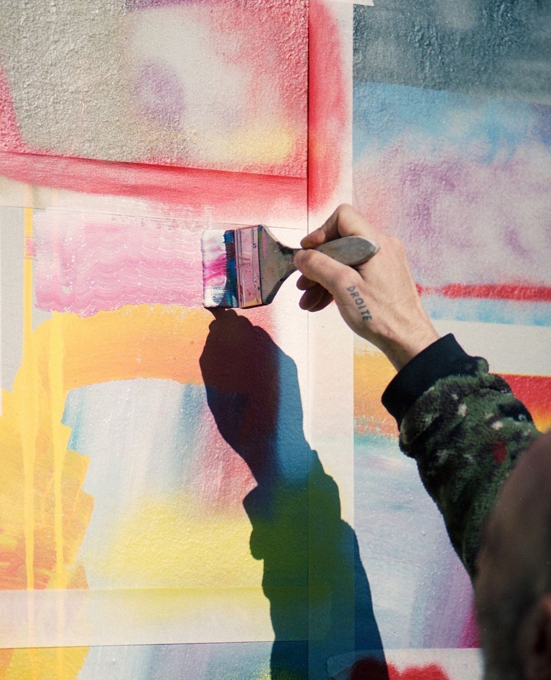 A hand with a tattoo holds a paint brush, painting colorful pastel shades onto a canvas, creating an abstract mural in daylight.