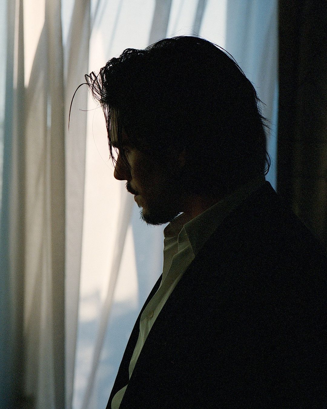 Portrait of a silhouetted adult male wearing a suit and white shirt, posing indoors near a window with natural light.