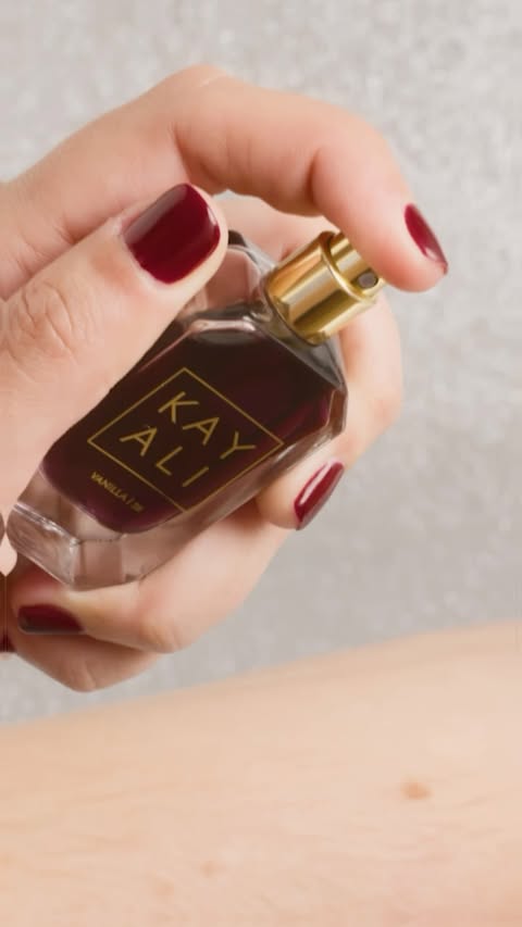 Woman sprays Kayali Vanilla 28 perfume on her arm. Close-up captures the bottle, hand, and arm against a neutral background. 