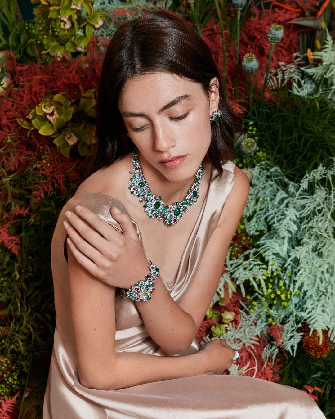 Elegant woman in champagne dress wearing emerald and diamond jewelry set against a vibrant floral background, seated indoors.