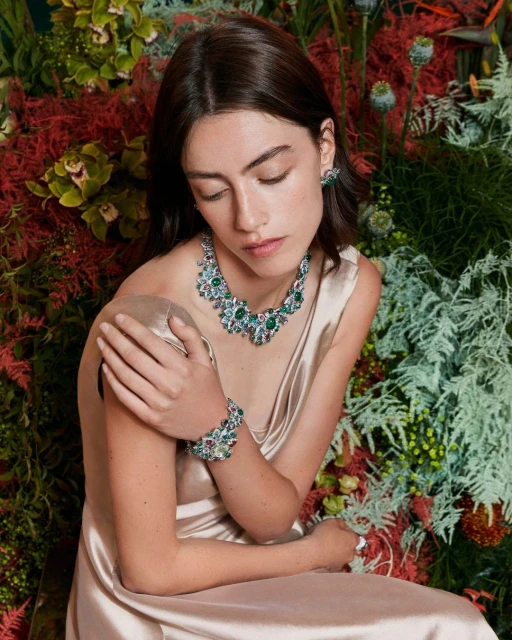 Elegant woman in champagne dress wearing emerald and diamond jewelry set against a vibrant floral background, seated indoors.