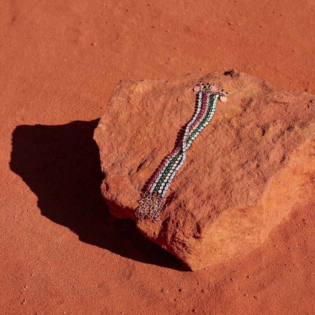 A close-up showcases a multi-strand, multi-colored bracelet resting on a textured rock surface, set against a reddish-brown background.