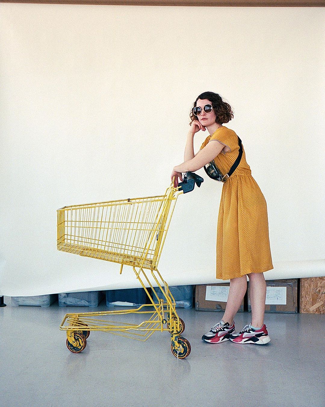 Woman in yellow dress leans on yellow shopping cart in a studio setting with neutral backdrop and a patterned fanny pack and sneakers.