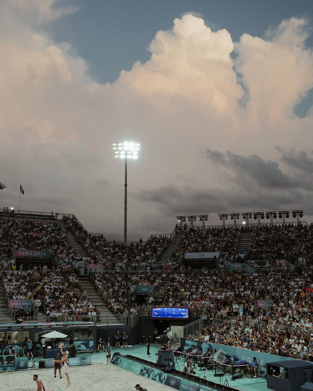 Crowd watches beach volleyball game on sand court under stadium lights and a cloudy sky at an outdoor arena.