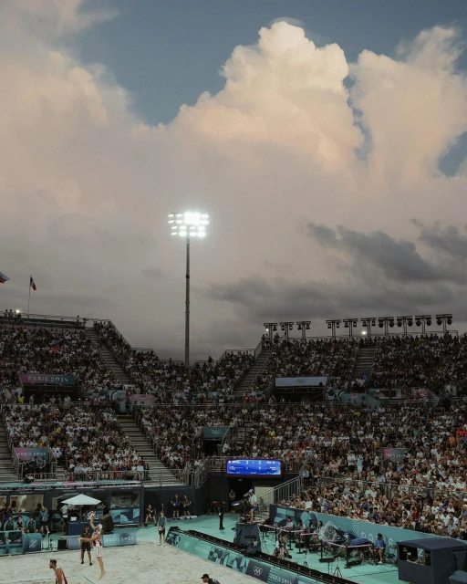 Crowd watches beach volleyball game on sand court under stadium lights and a cloudy sky at an outdoor arena.