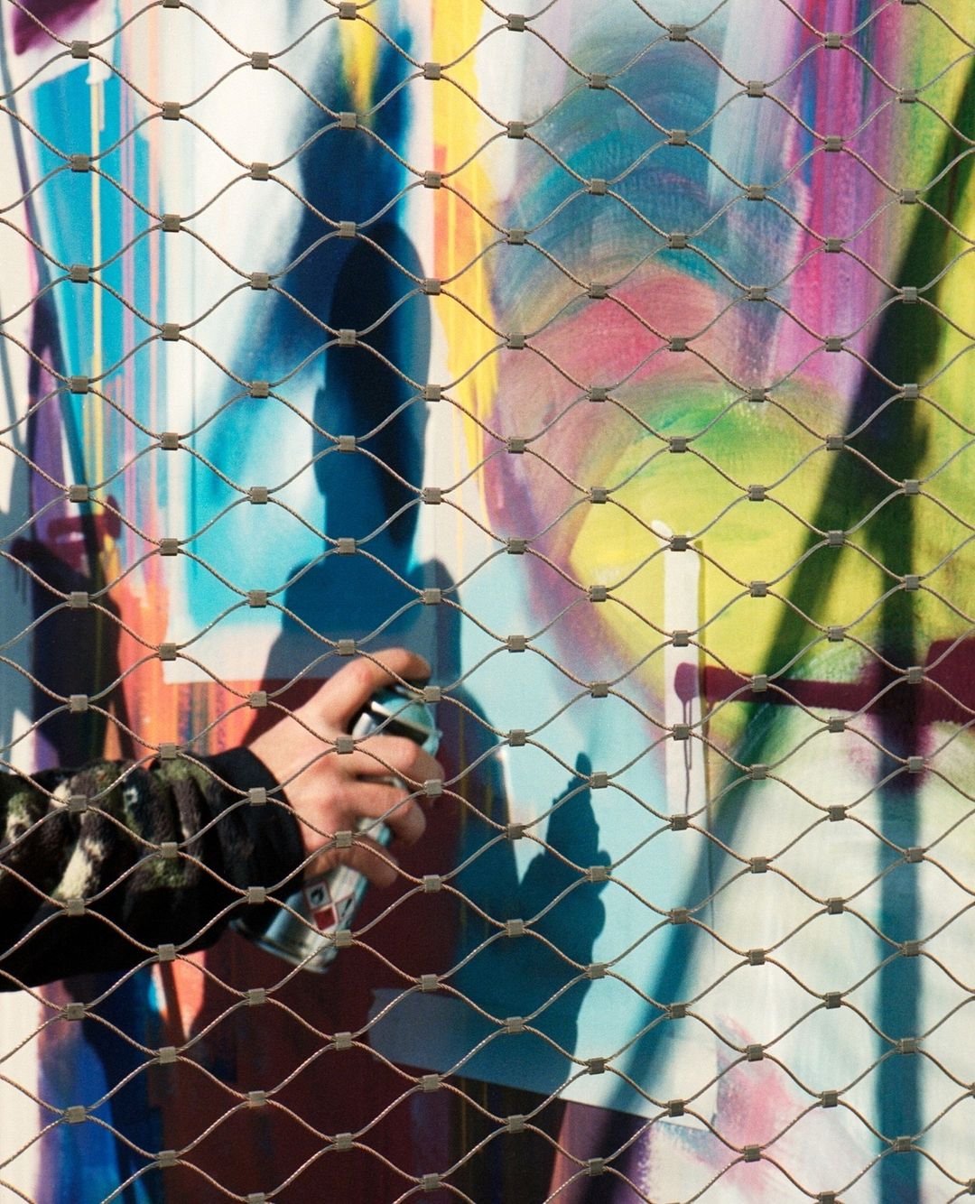 Person spraying graffiti art on a wall behind a wire mesh fence; arm, hand, spray can, urban art, and colorful backdrop visible.