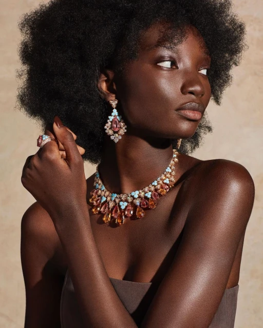 Close-up of a woman with dark skin wearing a brown strapless top and jewelry including a necklace, earrings, and ring against a neutral backdrop.