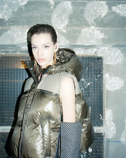 Fashion portrait of a woman wearing a hooded puffer vest and large gold earrings, set against a textured, tiled background.