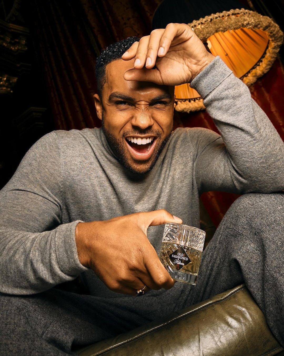 Close-up of a smiling man in grey loungewear holding a bottle of cologne indoors with a lamp visible in the background.