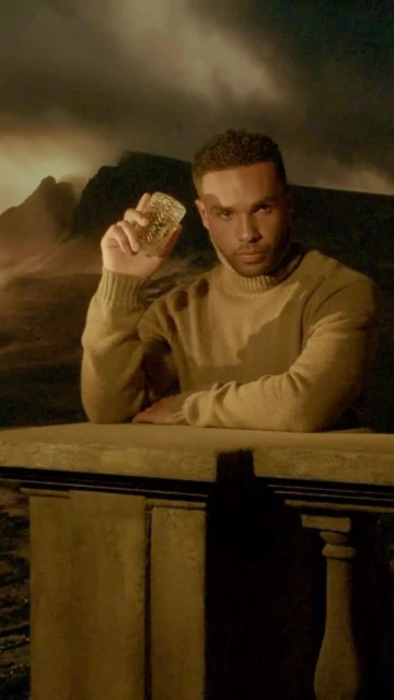 Man in a tan turtleneck sweater poses with a clear glass against a backdrop of mountains under a cloudy sky with subdued lighting.