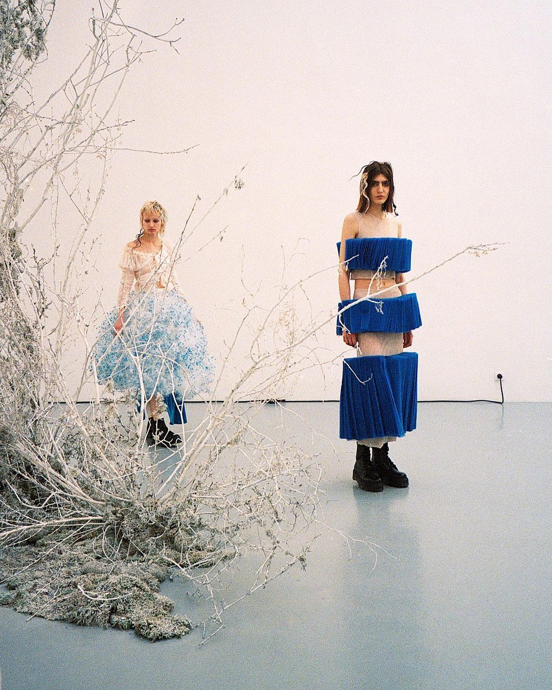 Two fashion models pose in an art studio against a white wall, one in a blue tiered dress, the other in a sheer dress by a white branch.