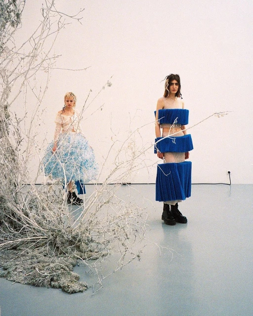 Two fashion models pose in an art studio against a white wall, one in a blue tiered dress, the other in a sheer dress by a white branch.