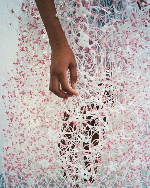An individual with an outstretched arm is draped in a dress made of white plastic tubes adorned with tiny pink spheres.