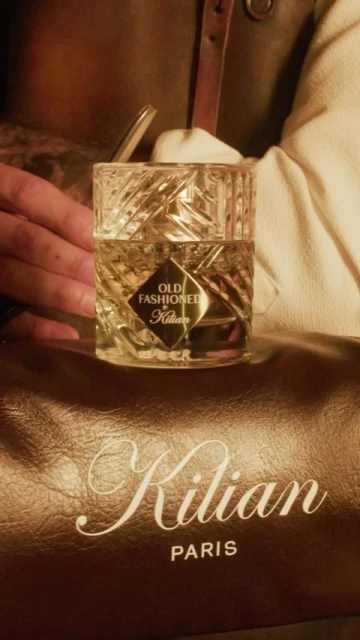 Crystal fragrance bottle of 'Old Fashioned' by Kilian on a leather bag, displayed by a hand, close-up product shot.