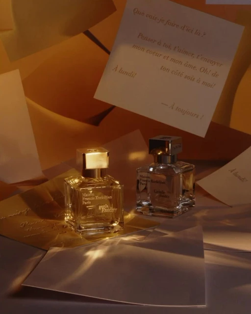 Two perfume bottles from Maison Francis Kurkdjian Paris on scattered paper with handwritten script. Gold and clear glass bottles, soft lighting.
