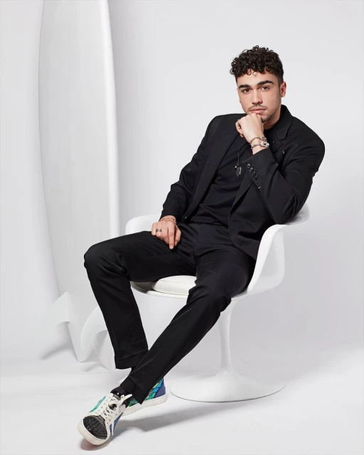 Man in a black suit poses in a chair with a surfboard in the background, wearing multicolored sneakers in a clean photo studio setting.