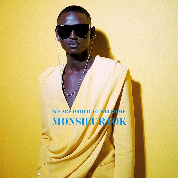 Black man wearing oversized sunglasses and a yellow draped suit jacket against a golden yellow background
