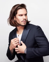 Man with long brown hair wearing a dark suit jacket adjusting his lapel against a plain white background