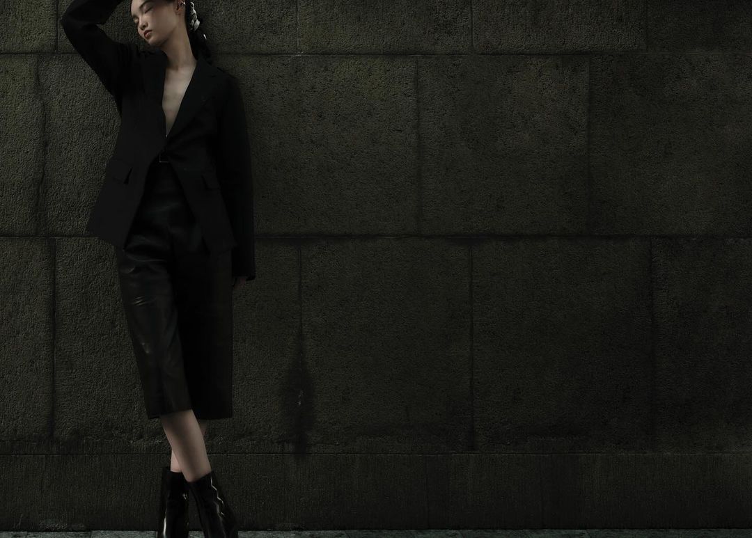 Portrait of a woman in a black blazer and culottes posing against a stone wall, creating a dramatic fashion editorial.