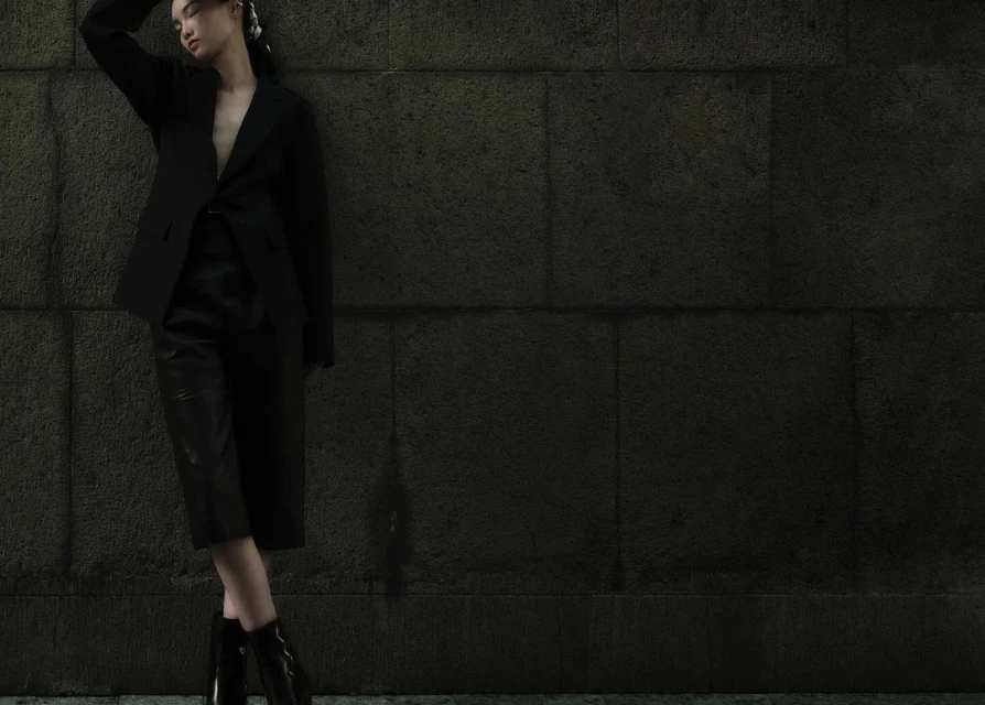 Portrait of a woman in a black blazer and culottes posing against a stone wall, creating a dramatic fashion editorial.