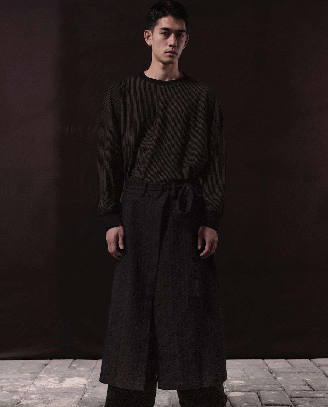 Portrait of a slender Asian man in a long-sleeve shirt and wrap pants, posing against a blurred dark brown background in studio lighting.