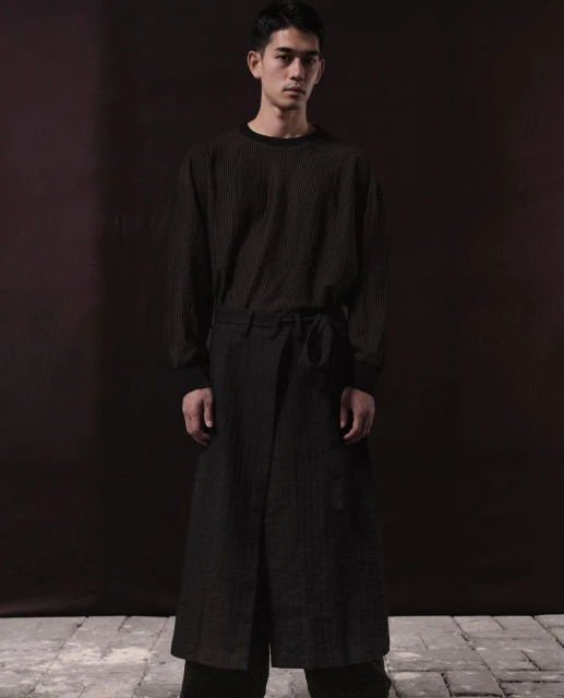 Portrait of a slender Asian man in a long-sleeve shirt and wrap pants, posing against a blurred dark brown background in studio lighting.