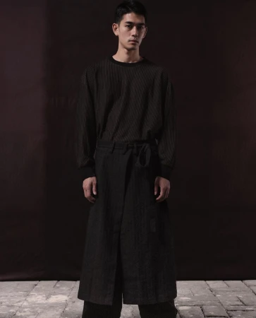 Portrait of a slender Asian man in a long-sleeve shirt and wrap pants, posing against a blurred dark brown background in studio lighting.