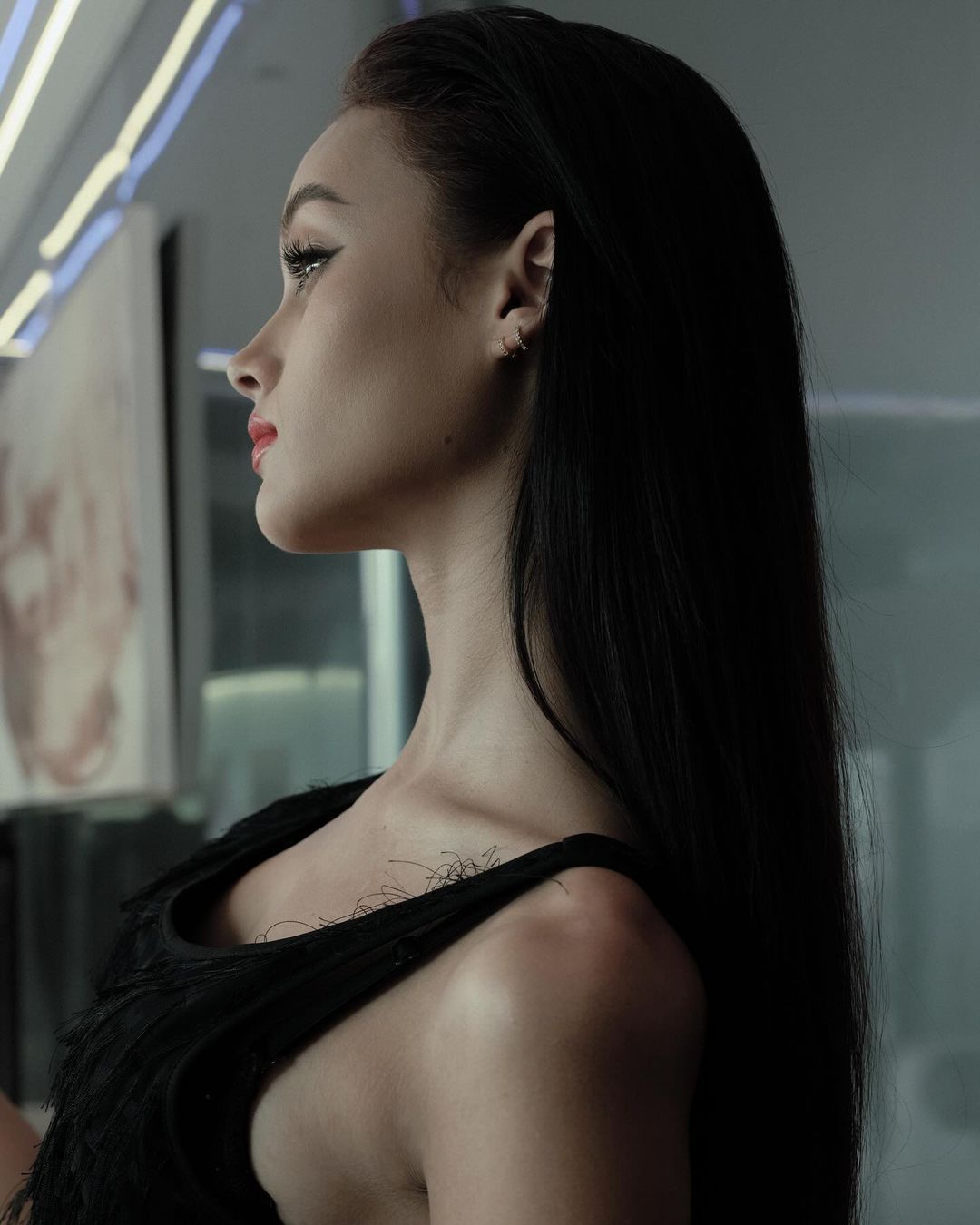 Profile portrait of a woman with long black hair wearing a black sleeveless top against an indoor interior background.