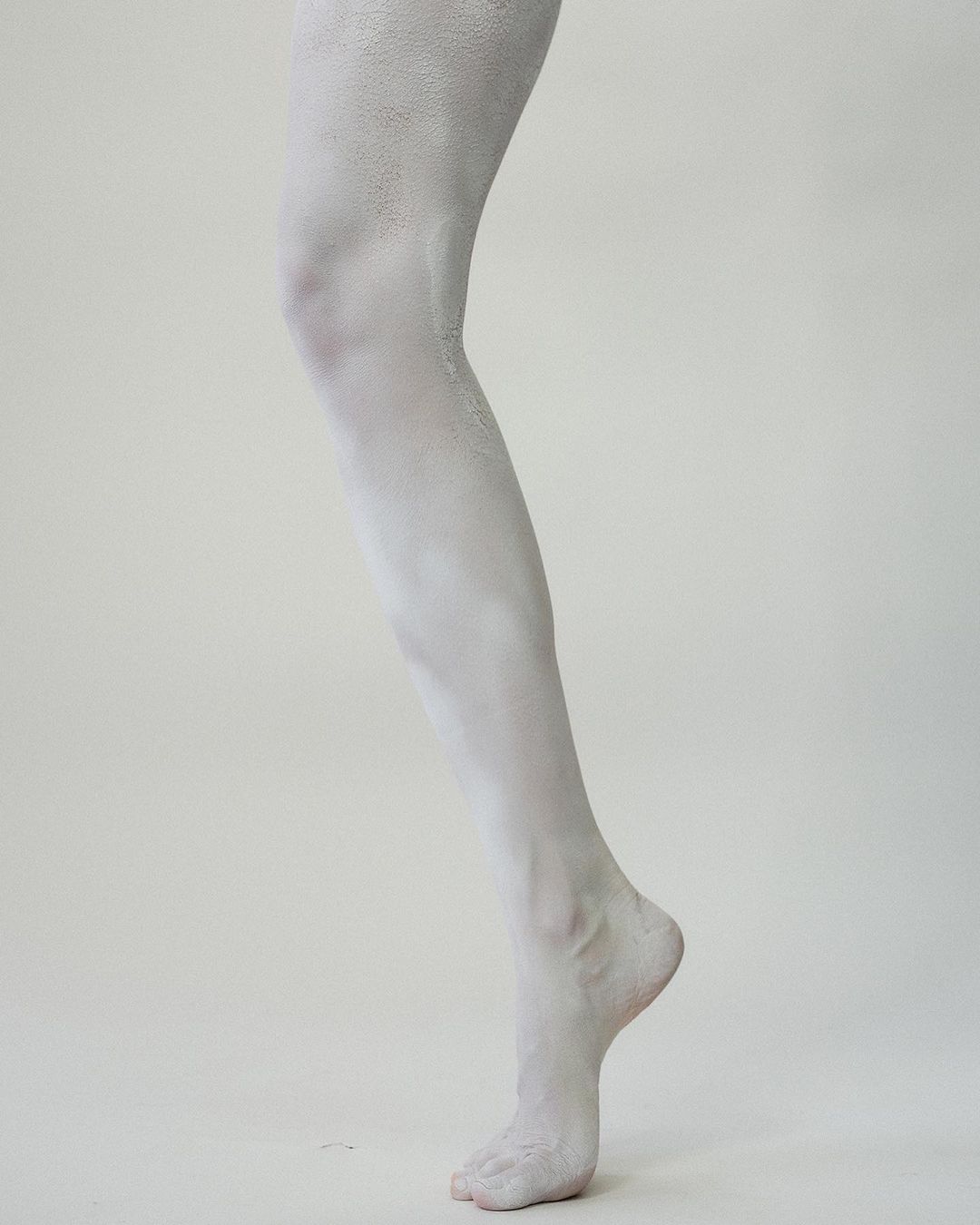 A close-up studio shot shows the lower half of an adult female covered in white paint standing against a neutral off-white background.