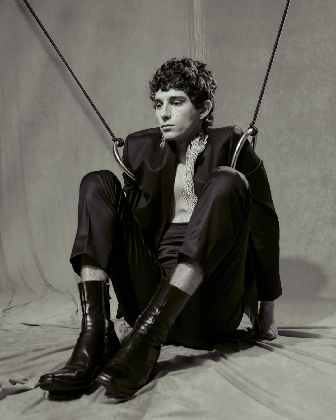Monochrome portrait of a young adult male model sitting in a studio wearing a black suit, bare chest, and boots with ring props.