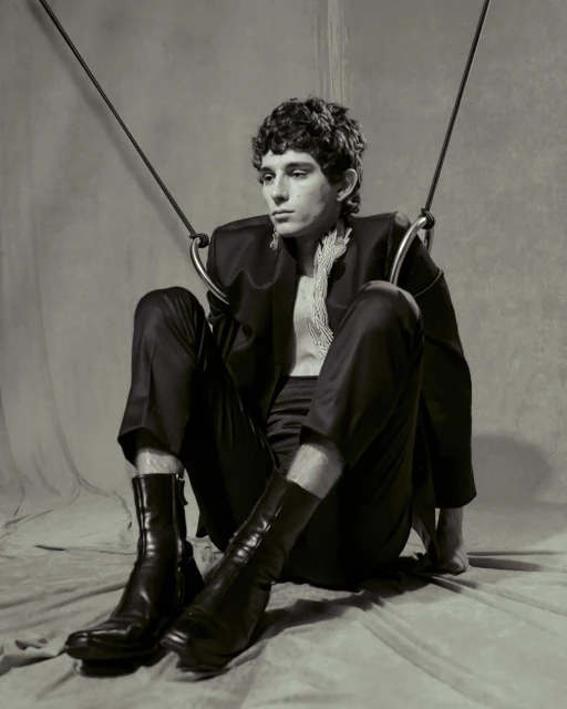 Monochrome portrait of a young adult male model sitting in a studio wearing a black suit, bare chest, and boots with ring props.