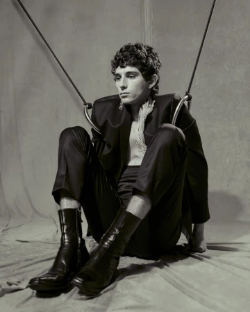 Monochrome portrait of a young adult male model sitting in a studio wearing a black suit, bare chest, and boots with ring props.