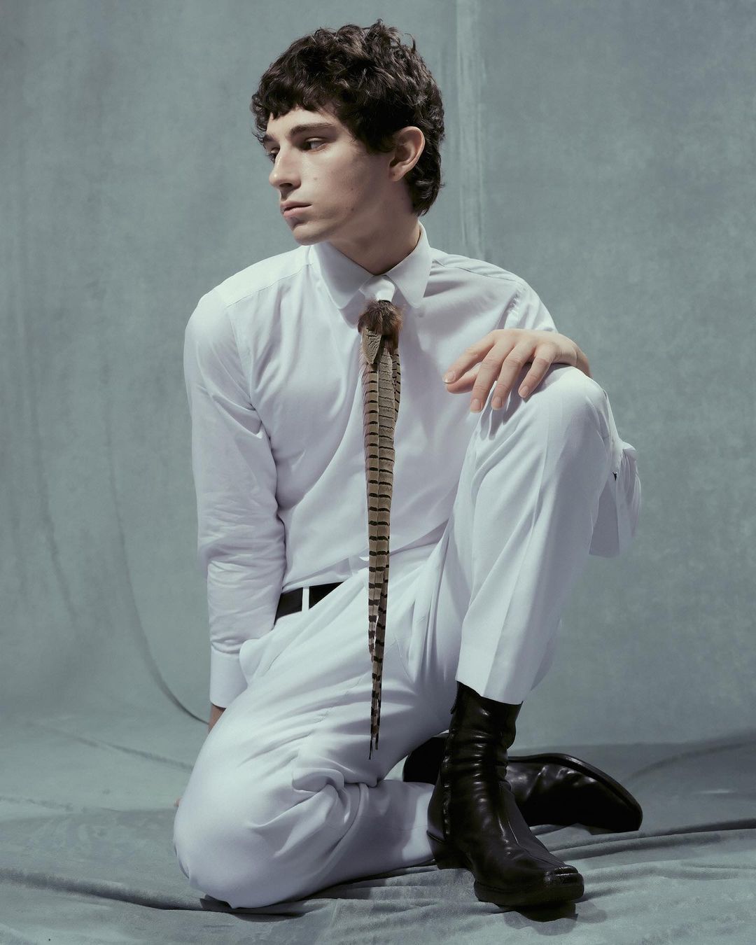 Adult male with curly hair poses in white shirt and trousers, wearing a feather tie and black boots, set against a grey studio backdrop.