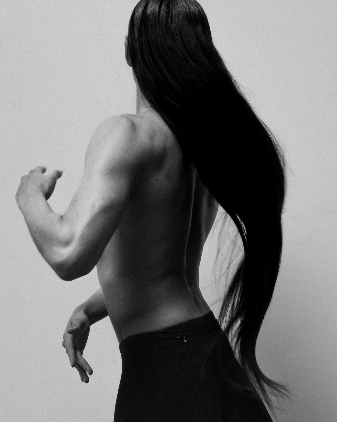 Striking black and white studio shot of a shirtless person with long dark hair, back to camera, posed against a bright white backdrop.