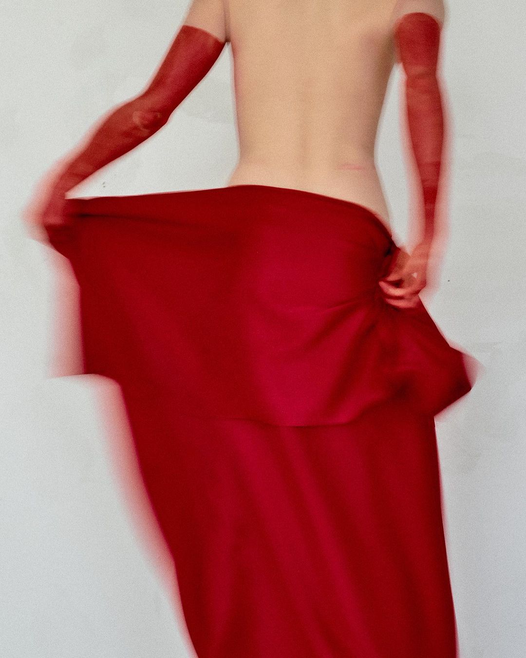 Back view of bare-backed adult with long red opera gloves and flowing red skirt in motion blur, against a plain white backdrop.