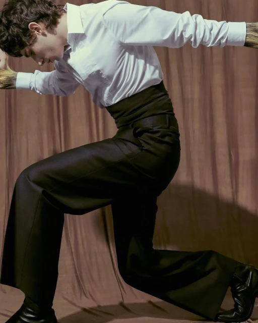 Fashionable adult in a white dress shirt and black slacks strikes a dynamic pose against a neutral backdrop in a studio shot.