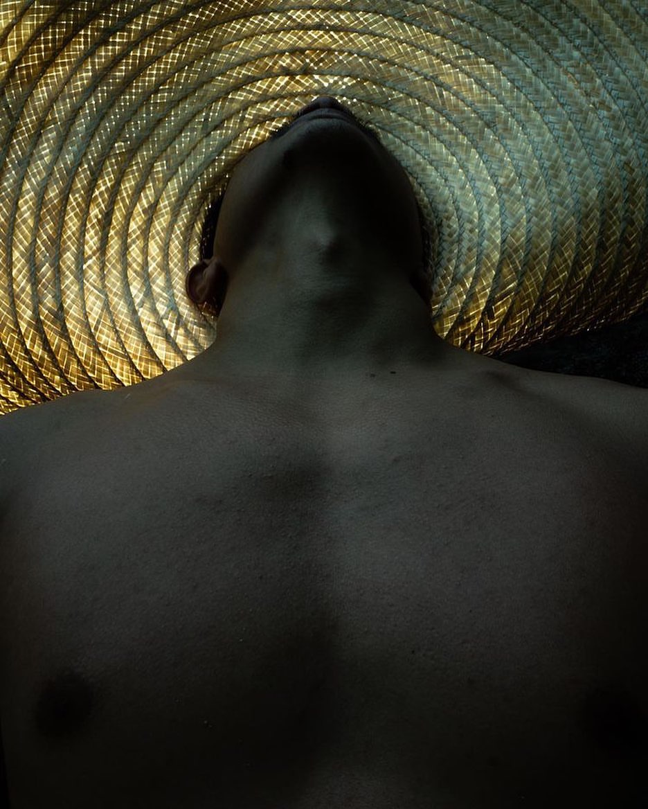 Low-angle shot of a shirtless man wearing a straw hat with a spiral pattern, with dramatic lighting and a focus on texture and form.