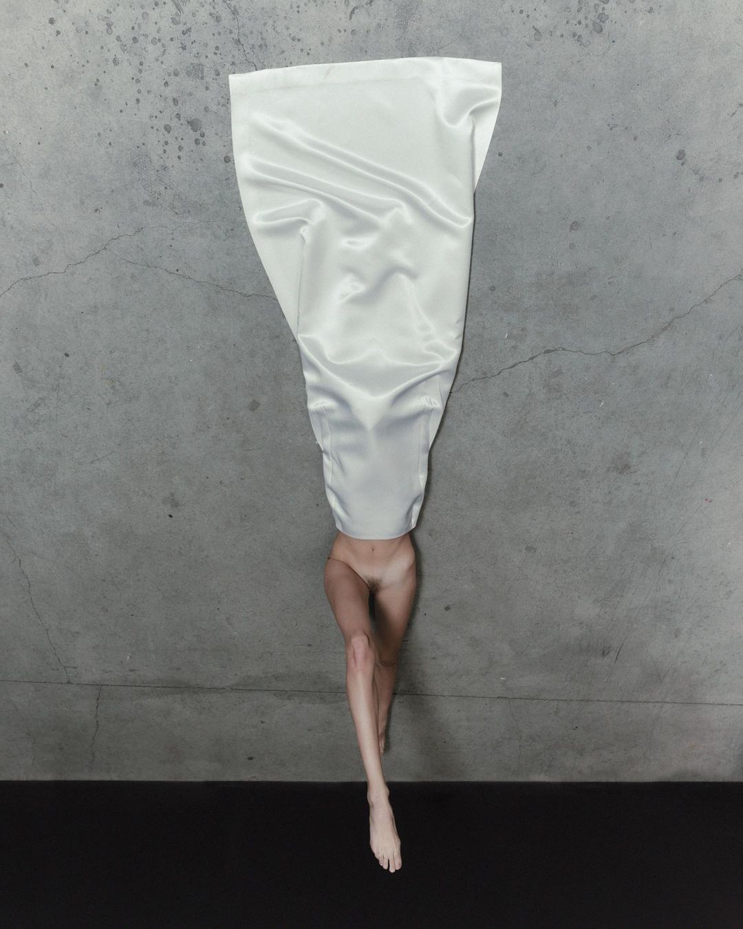 Surreal image of a woman's legs and torso emerging from draped white fabric against a gray concrete backdrop on a black surface.