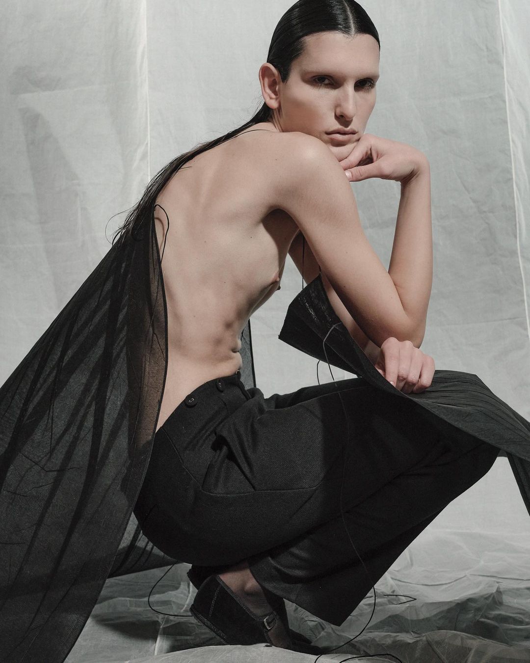 Person in dark trousers and sheer garment, crouching in a studio with neutral background, fashion or fine art photography. 