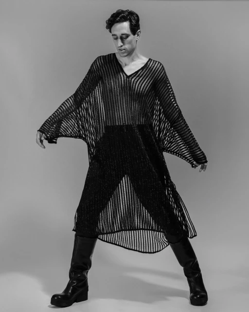 Black and white portrait of a slender adult man wearing a sheer, striped black tunic, fitted black trousers, and black leather boots in a studio.