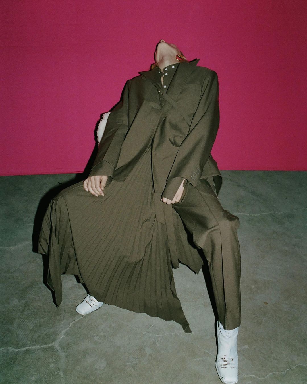 Woman in oversized olive-green suit and skirt poses in a chair against a pink background, wearing white shoes, fashion-forward.