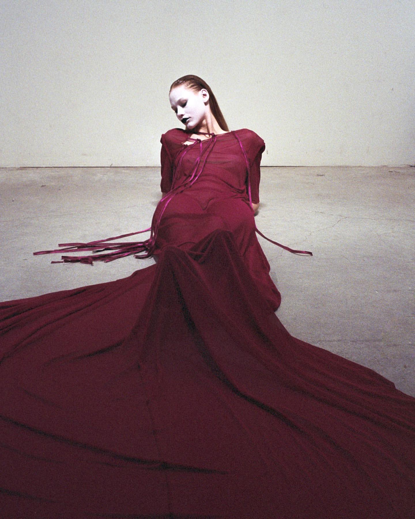 Fashion shot of a model sitting in a studio wearing a long, dark-red dress with train, and heavy white face makeup, looking downward.