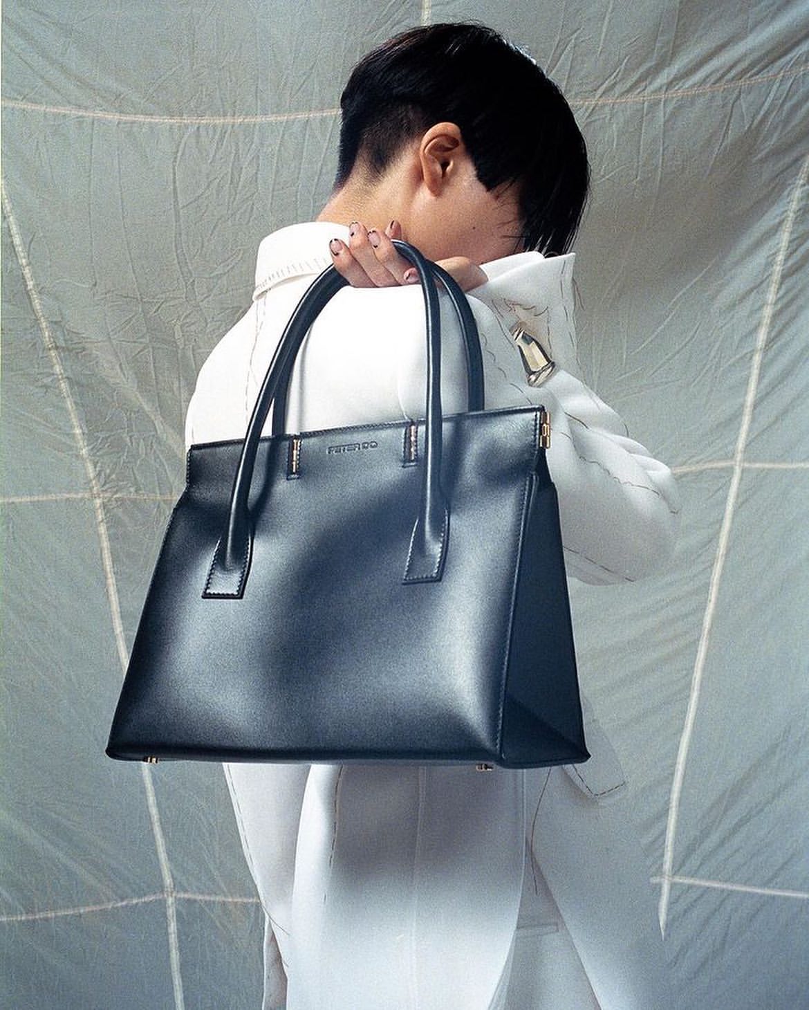 Adult holds a black leather tote bag in front of a white textured backdrop in a studio setting, showcasing fashion accessory.