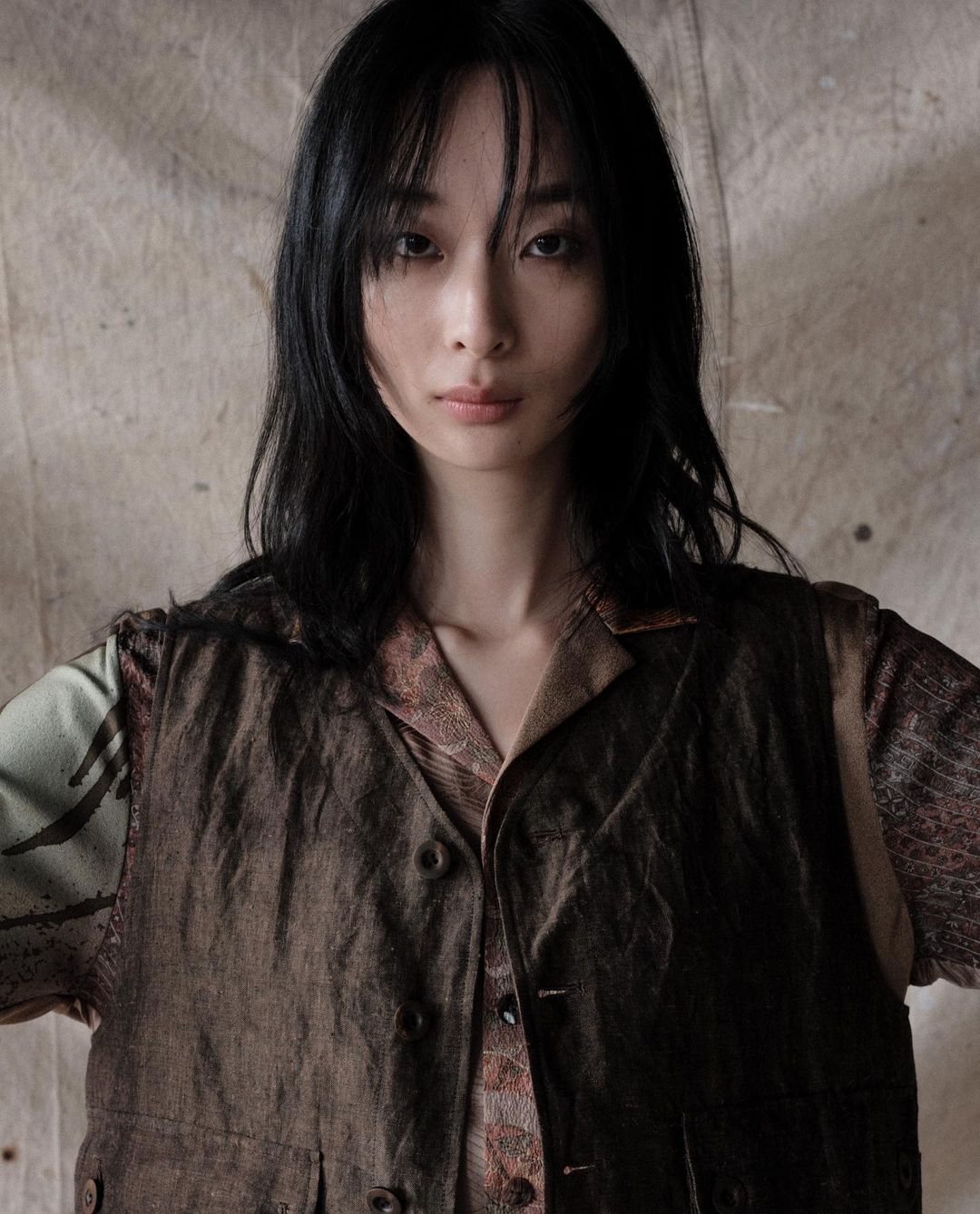 Portrait of a young Asian woman in a brown vest and patterned shirt against a textured backdrop, looking directly at the camera.